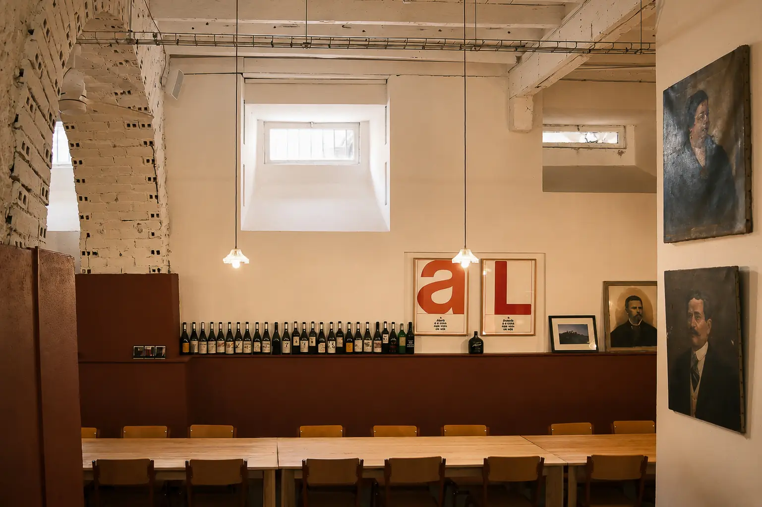 Mesa corrida posta na sala principal do Couvert Lisboa, com pé-direito alto, parede de tijolo pintada e garrafas de vinho alinhadas.
