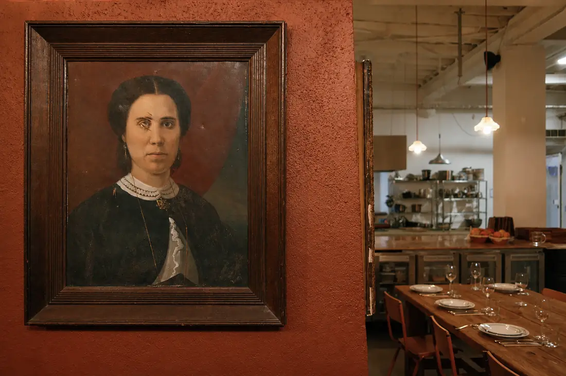 Retrato a óleo sobre a parede de tijolo, com a mesa posta ao lado.